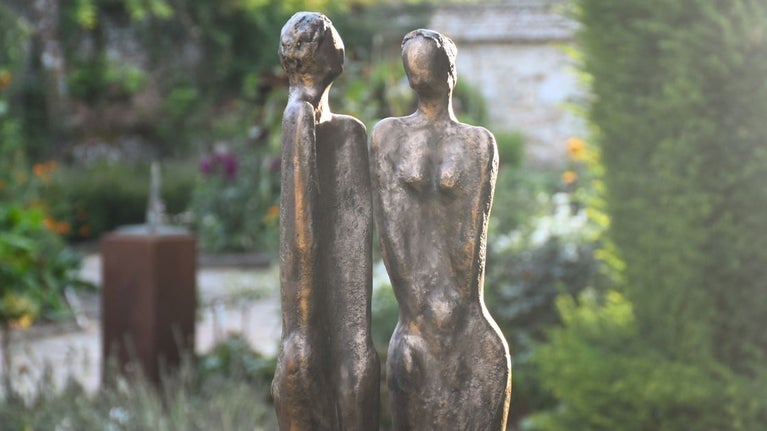 A close up of two figurines, a man and a woman against a blurred background of clipped green topiary and bright coloured flowers